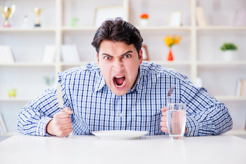 Hangry.jpg Ein wütender Mann hält ein Messer und eine Gabel über einem leeren Teller, während er an einem Tisch sitzt.