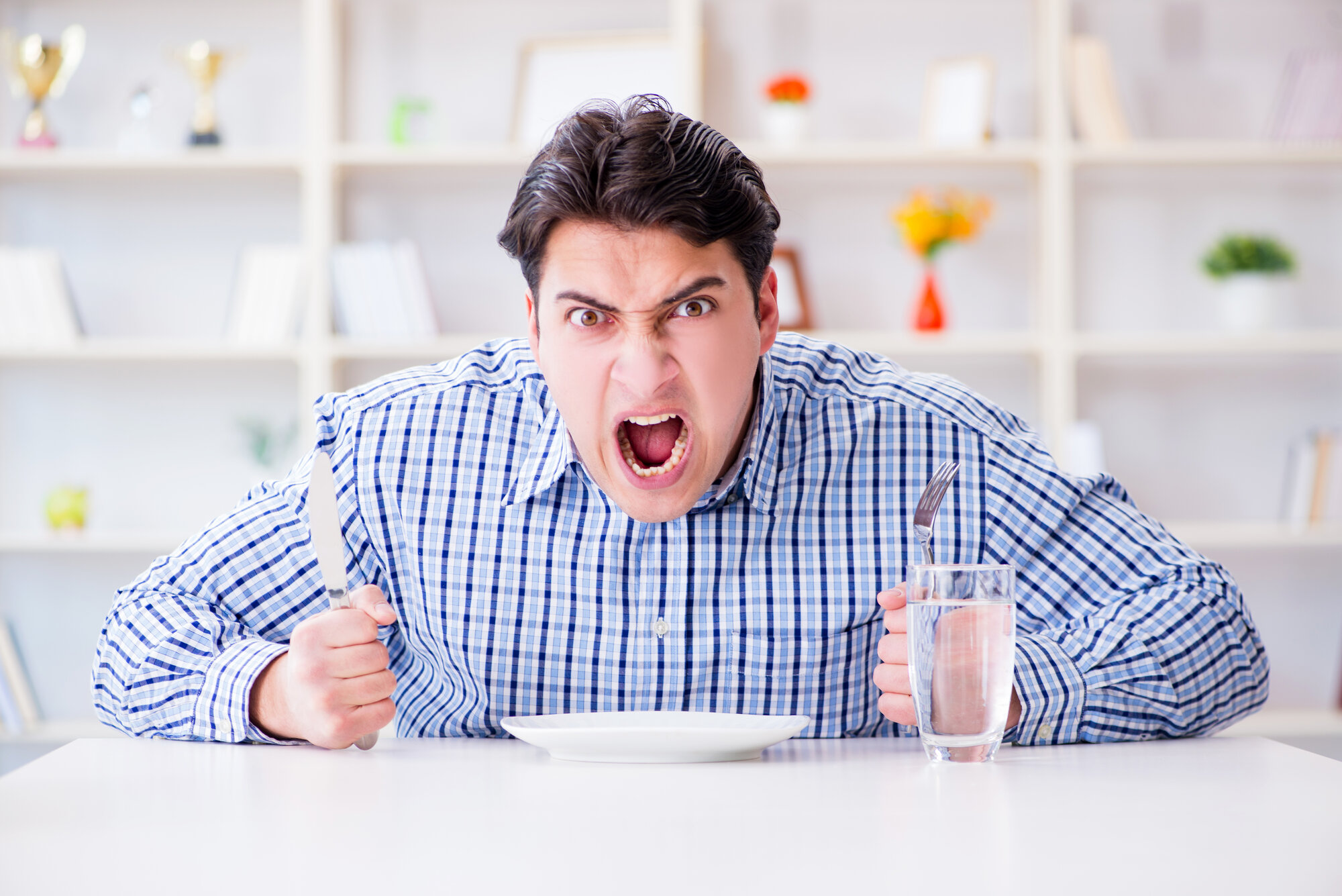 Hangry.jpg Ein wütender Mann hält ein Messer und eine Gabel über einem leeren Teller, während er an einem Tisch sitzt.