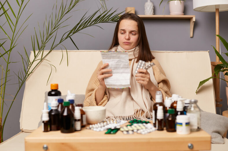 selbstmedikation.jpg Eine kranke Frau sitzt auf einem Sofa, umgeben von verschiedenen Medikamenten und hält einen Zettel sowie eine Medikamentenpackung in der Hand.