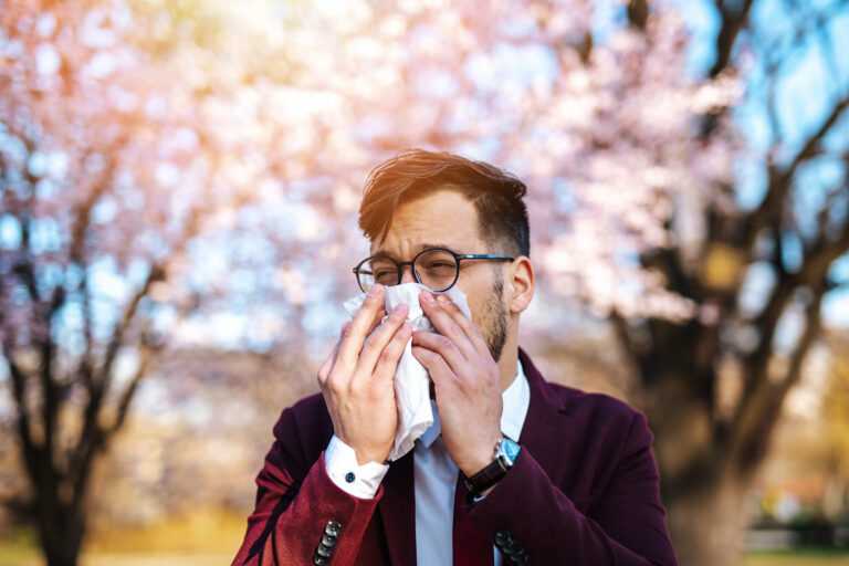 Ein Mann in einem Anzug benutzt ein Taschentuch, um sich die Nase zu putzen, vor blühenden Bäumen im Hintergrund.