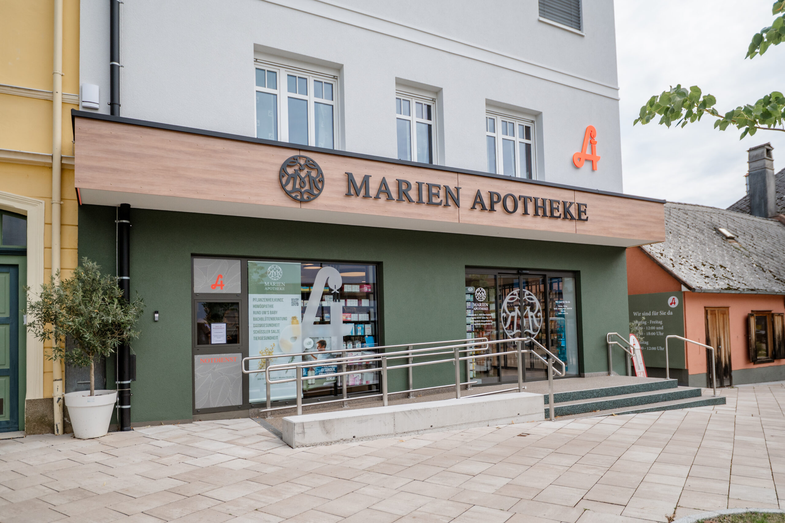Außenansicht der „Marien Apotheke“ in einem modernen Gebäude mit grünem Erdgeschoss und hellem Obergeschoss. Über dem Eingang steht der Schriftzug „MARIEN APOTHEKE“ auf einer Holzverkleidung, daneben ein rotes Apothekenzeichen. Der Eingang ist barrierefrei mit Rampe und Treppe ausgestattet.