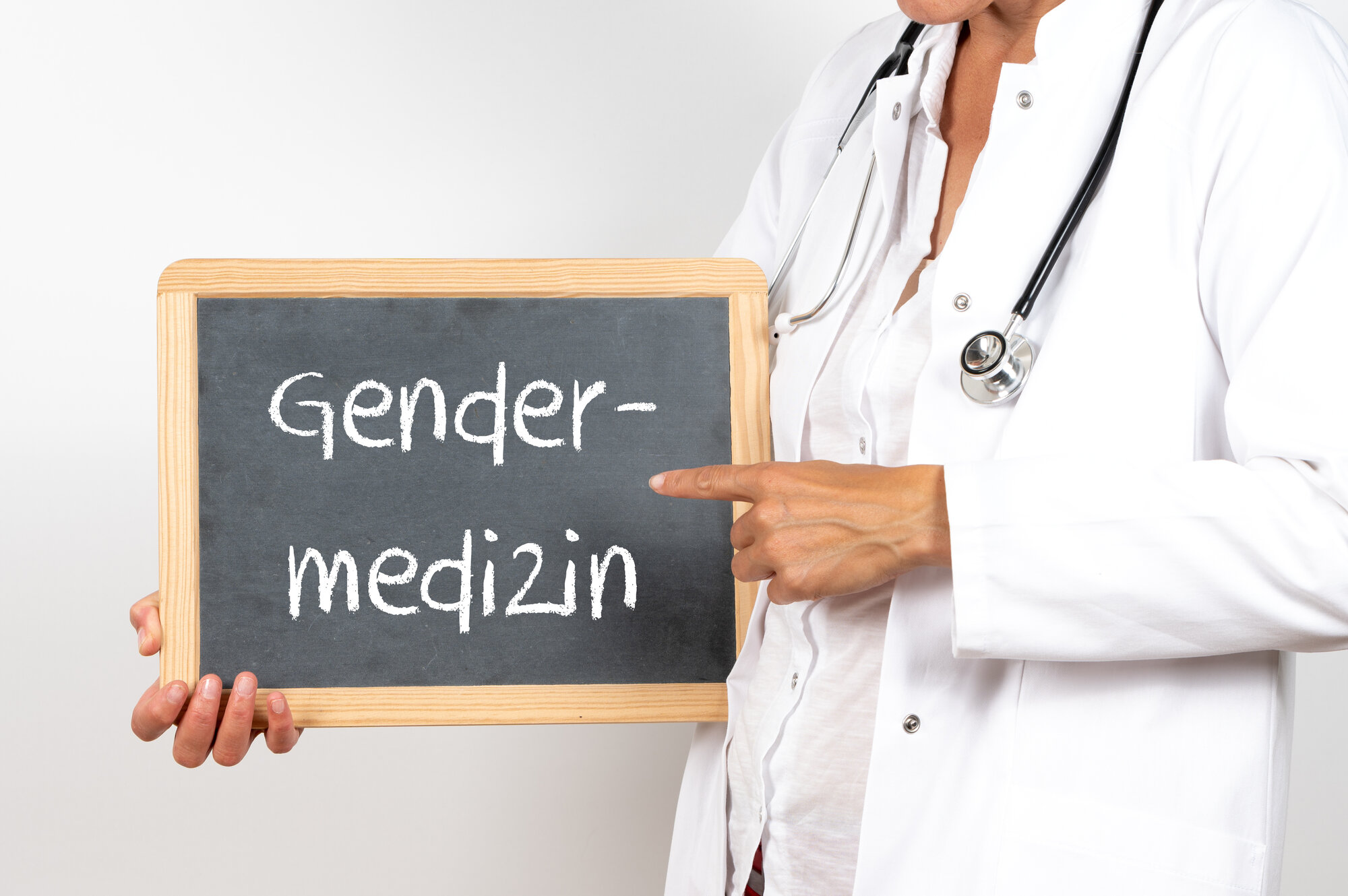 Gendermedizin.jpeg Eine Person in Arztkleidung hält eine Tafel mit der Aufschrift "Gendermedizin".